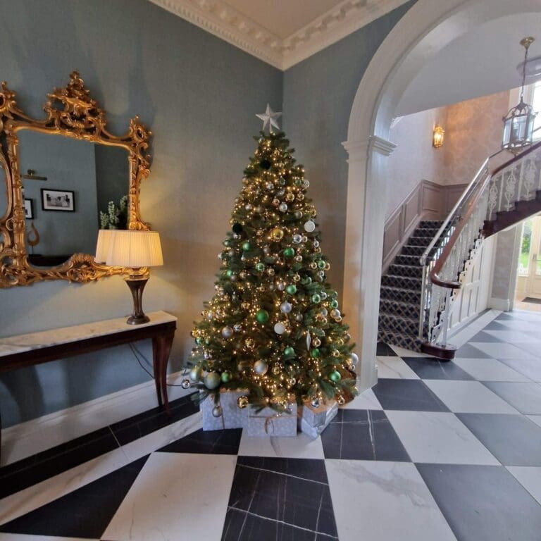 Christmas tree with warm lights in a hallway with a chequered floor, gilt mirror and staircase
