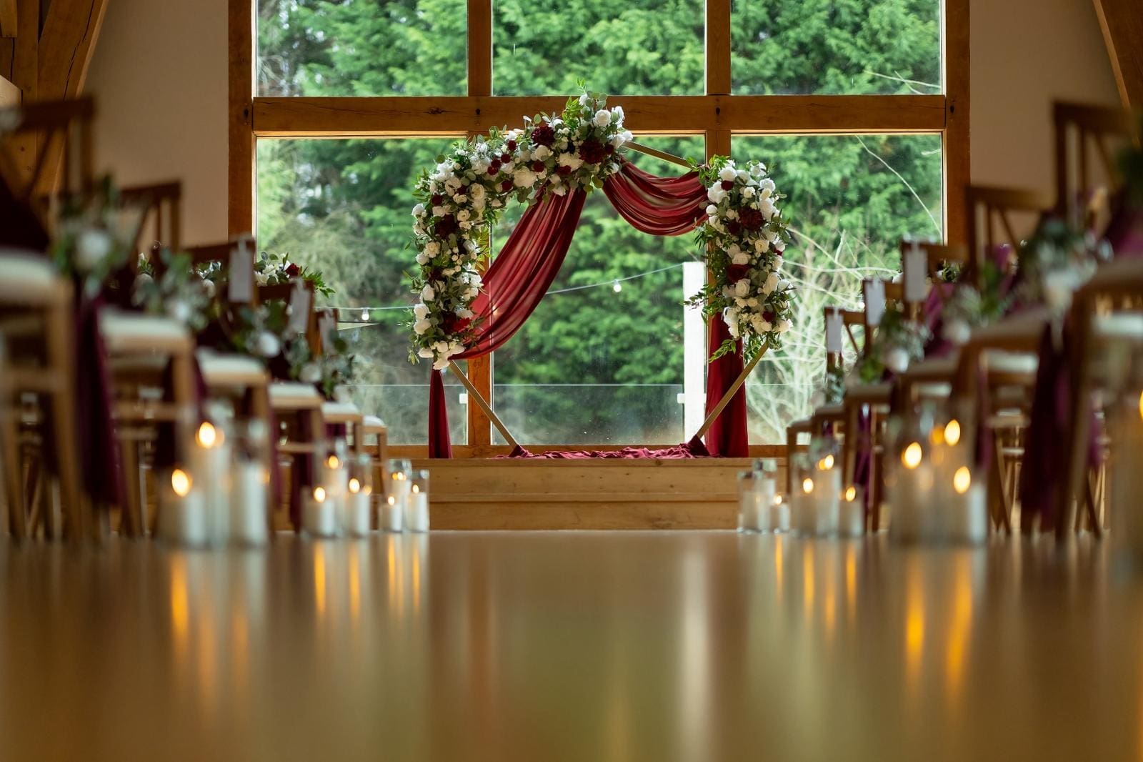 Hexagon wedding arch with burgundy draping and white florals at the end of a candlelit aisle at The Mill Barns