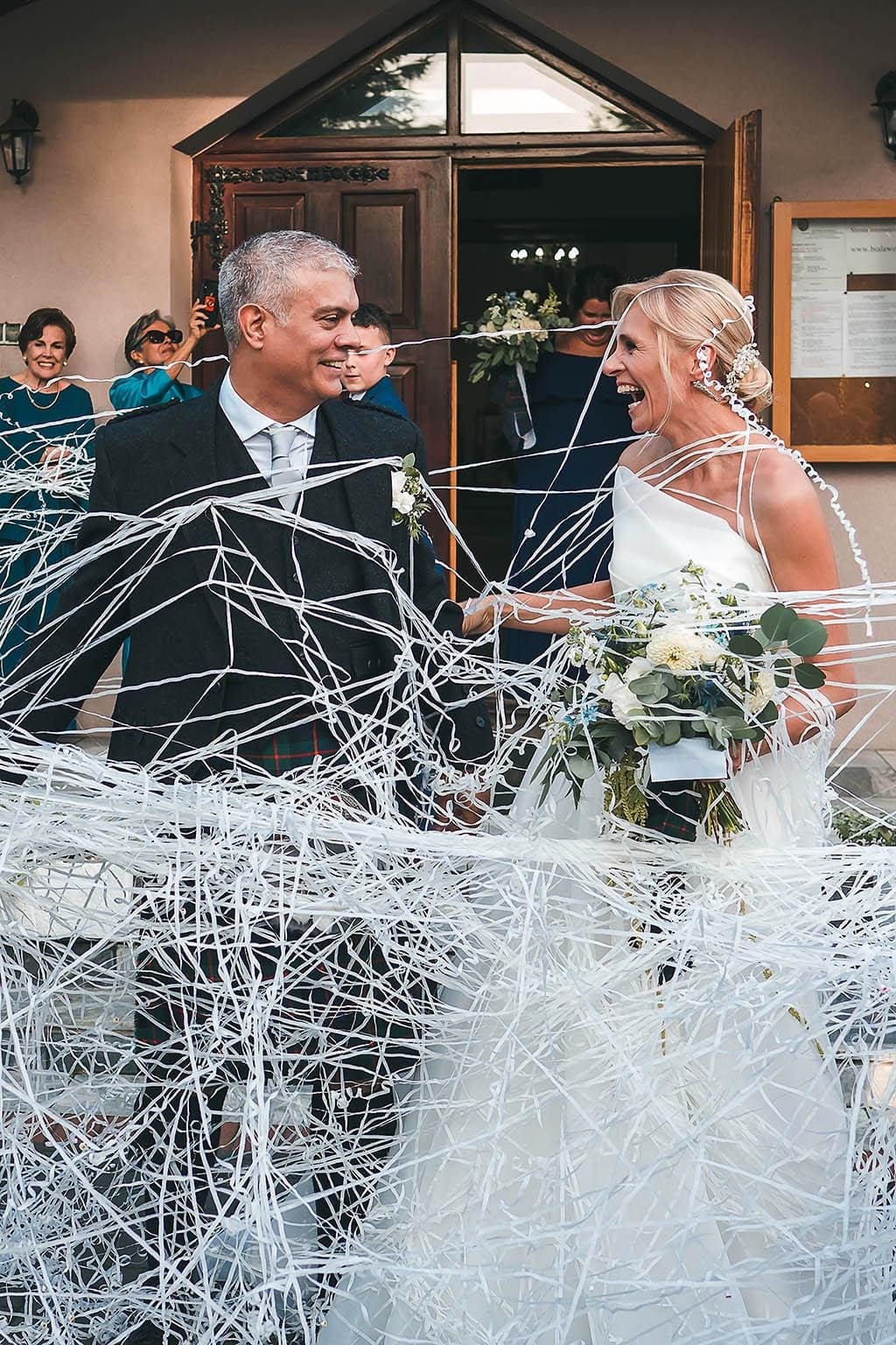 Bride and groom laughing together covered in paper streamer confetti as they leave their wedding ceremony