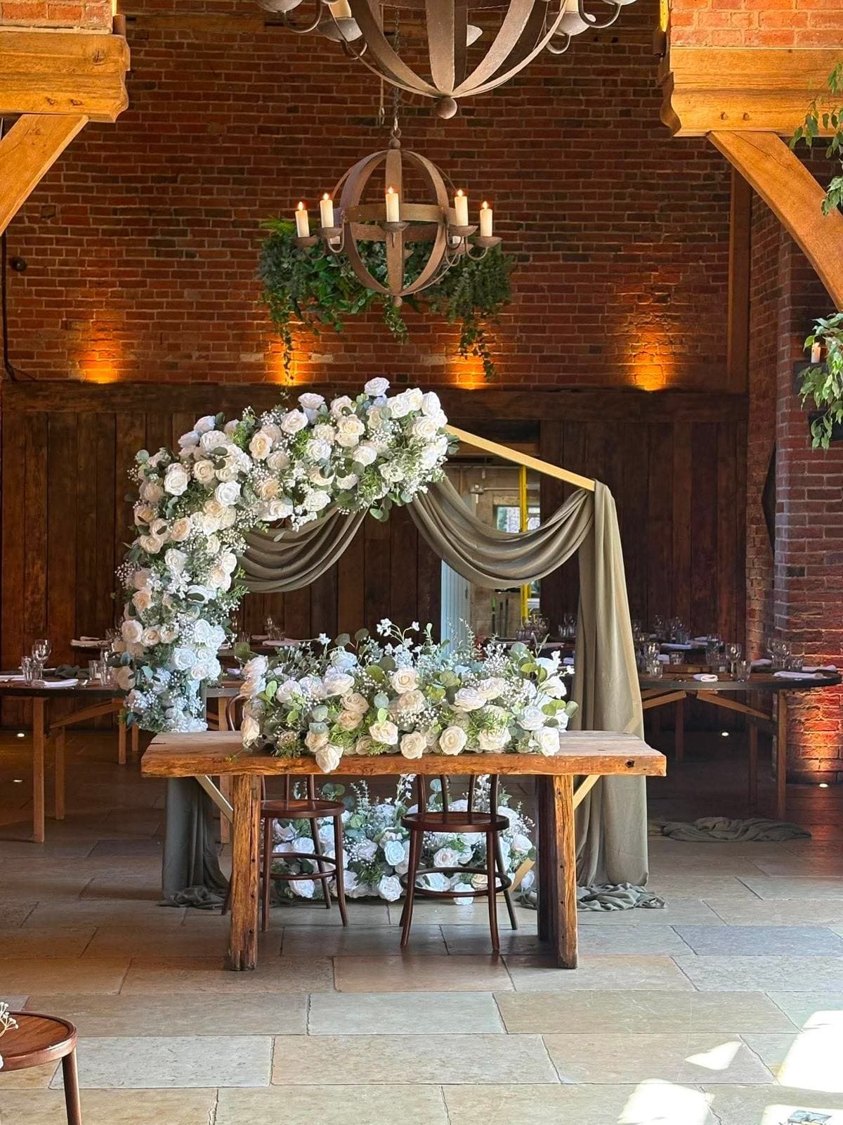 Isla Range white rose broken arch with sage drapes over a rustic top table with matching flowers, in a brick barn